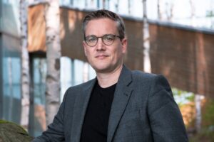 Sebastian Bluhm Portraitbild mit grauem Sakko und schwarzen T-Shirt vor Waldhintergrund