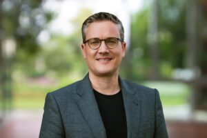 Sebastian Bluhm mit Sakko und schwarzem T-Shirt als Portraitbild vor grünem Hintergrund